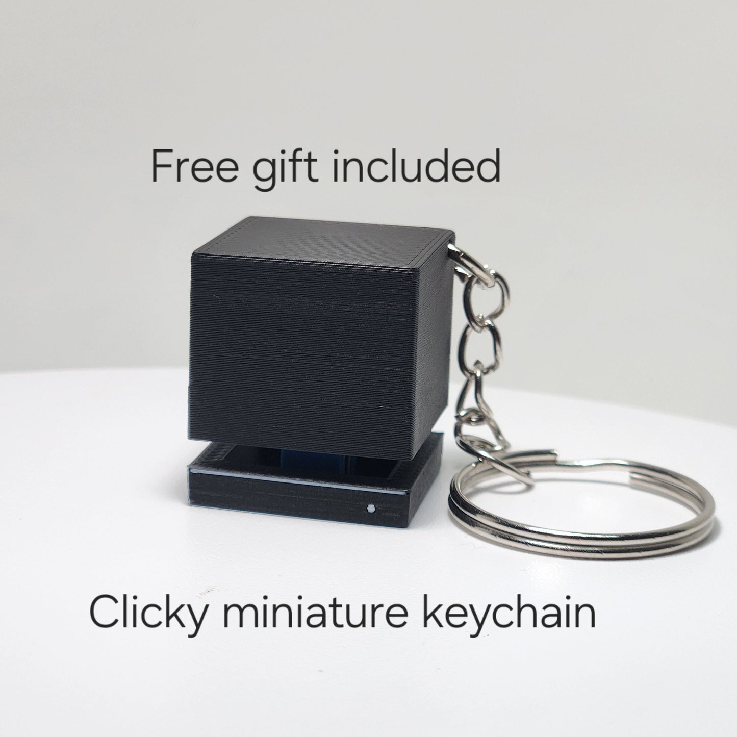 Black clicky miniature keychain on a white surface with text indicating a free gift offer.